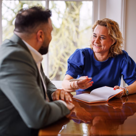 Man and woman in a meeting, with the woman wearing a blue top taking notes while smiling during the conversation.