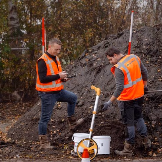 Two people wearing reflective vests inspecting a pile of soil, pointing at white buckets while one person takes notes on an iPad.