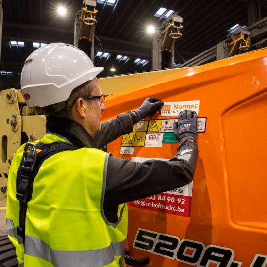 Industrial worker wearing high-visibility safety clothing and a hard hat applies or inspects a certification label on heavy machinery in a manufacturing facility, highlighting equipment inspections, machinery certification, regulatory compliance, workplace safety, and quality assurance for industrial equipment.