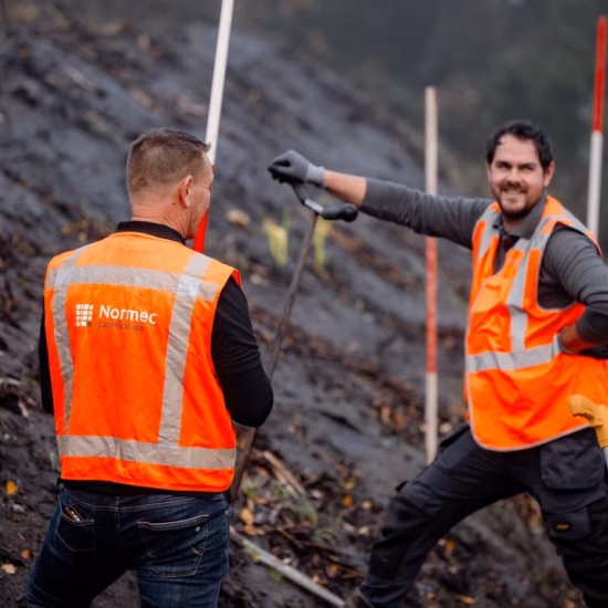 Twee medewerkers van Normec werken op een hellend terrein naast een infrastructureel project. Met meetapparatuur en veiligheidskleding voeren zij veldmetingen en inspectiewerkzaamheden uit. Op de achtergrond zijn markeringsstokken zichtbaar langs het talud.