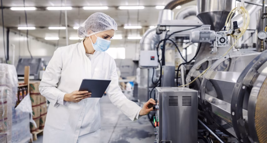 A quality control worker inspects an industrial production line at a food processing plant using a tablet and personal protective equipment.
