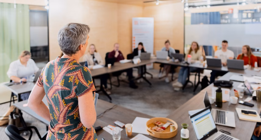 Deelnemers nemen deel aan een bijeenkomst of training in een vergaderruimte, terwijl een spreker voor de groep presenteert.