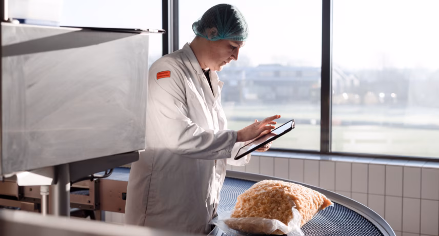 An employee wearing protective work clothing inspects a bread product using a tablet next to a conveyor belt in a production environment.