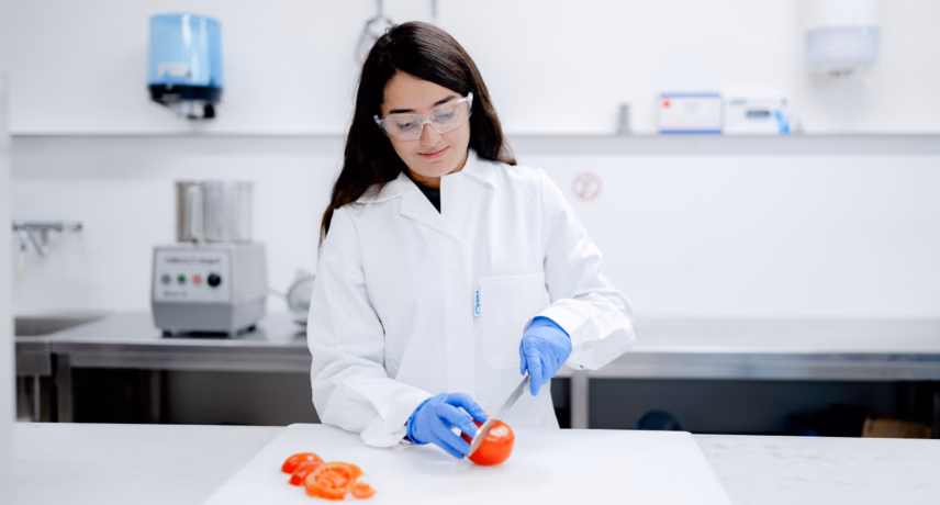 Ein Laborant in Schutzkleidung führt im Labor eine Qualitätskontrolle an einer Tomate durch.