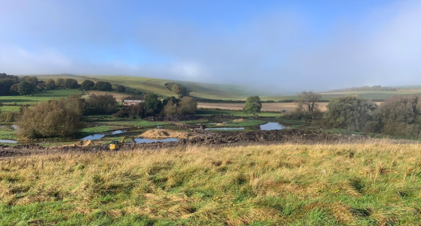 A rolling grassland landscape with fields, a few bodies of water, and scattered buildings under a slightly cloudy sky.