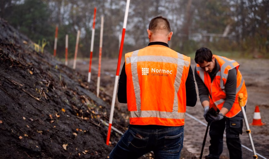 Employees wearing safety vests are conducting fieldwork at a soil investigation site using measuring equipment.