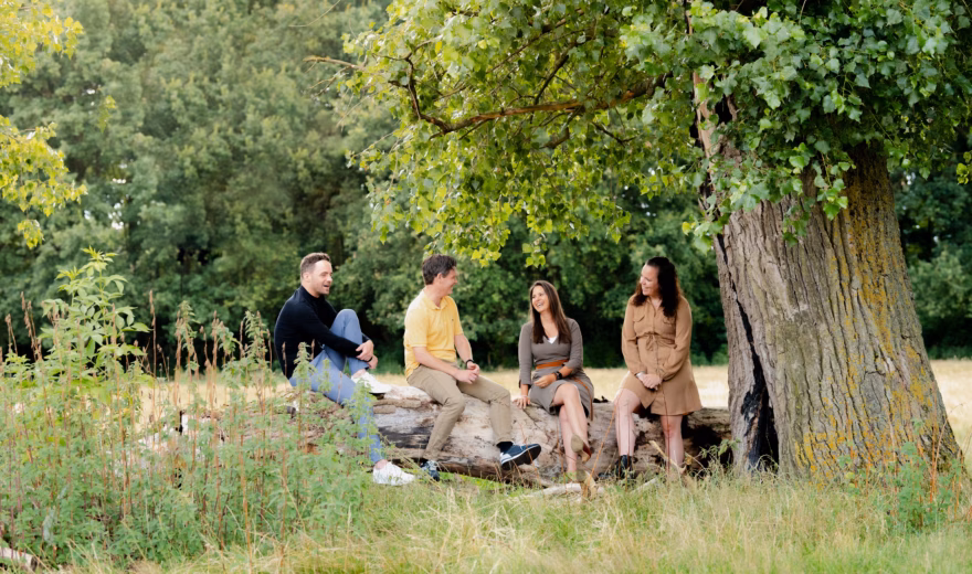 Groep mensen zit samen op boomstammen onder een grote boom in een groene natuurlijke omgeving.