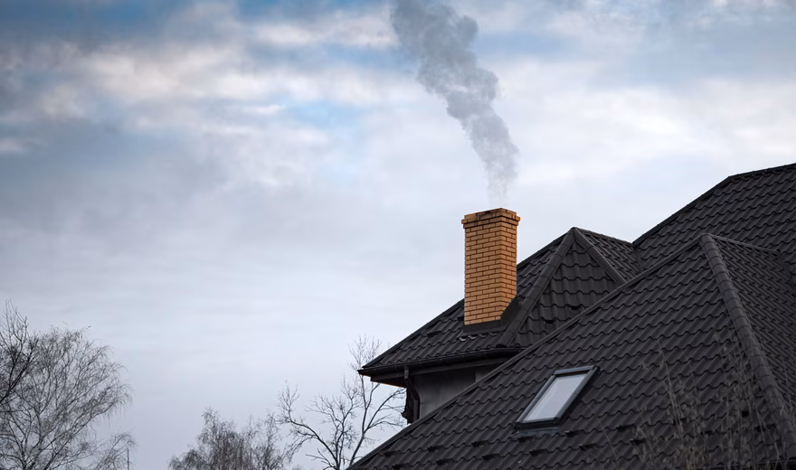Een schoorsteen op het dak van een woning stoot zichtbare rook uit tegen een bewolkte lucht. De afbeelding illustreert het gebruik van een houtkachel, open haard of ander verwarmingssysteem waarbij verbrandingsgassen via de schoorsteen worden afgevoerd.
