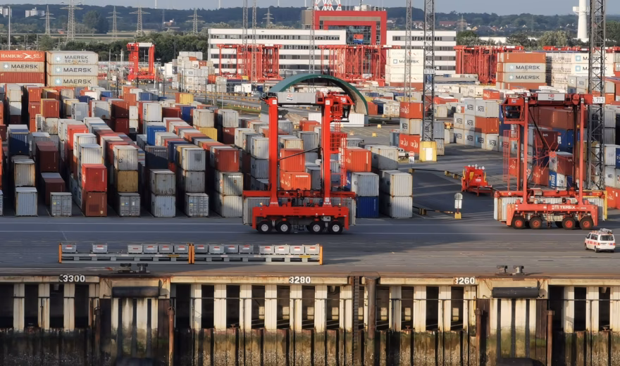 Overzicht van een grote containerterminal waar honderden zeecontainers zijn opgeslagen in lange rijen. Rode portaalkranen en overslagmaterieel verplaatsen containers op het terrein langs de kade, als onderdeel van de logistieke en internationale goederenstromen.