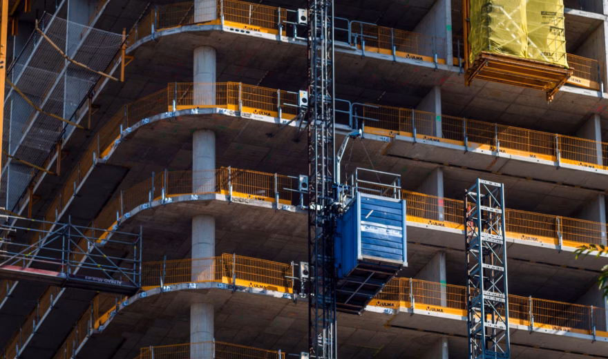 Plusieurs étages d’un immeuble de grande hauteur en béton sont visibles pendant la phase de gros œuvre. Un ascenseur de chantier longe la façade pour assurer le transport vertical des matériaux et du personnel. Aux étages supérieurs, une charge de chantier emballée est hissée, tandis que des barrières de sécurité ont été installées le long des bords des planchers.