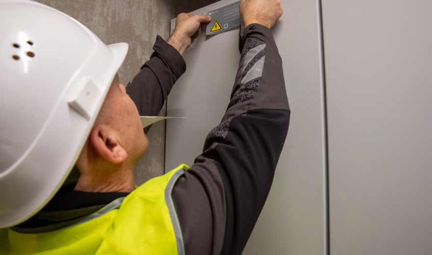 Een medewerker in persoonlijke beschermingsmiddelen brengt een identificatie- of waarschuwingslabel aan op een elektrische schakelkast. Op de kast is een waarschuwingssymbool voor elektrische spanning zichtbaar. De afbeelding laat werkzaamheden zien op het gebied van elektrotechniek, installatiebeheer en veiligheid.