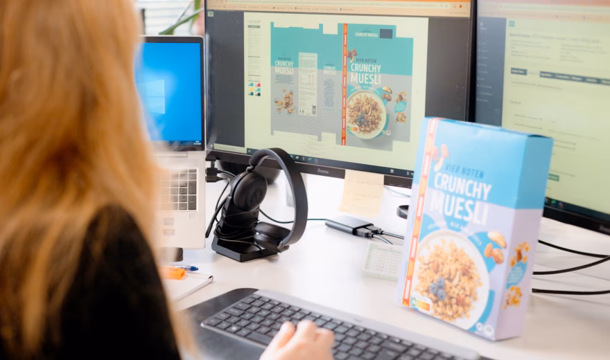 Medewerker werkt aan productverpakking op een computerwerkplek met meerdere monitoren.