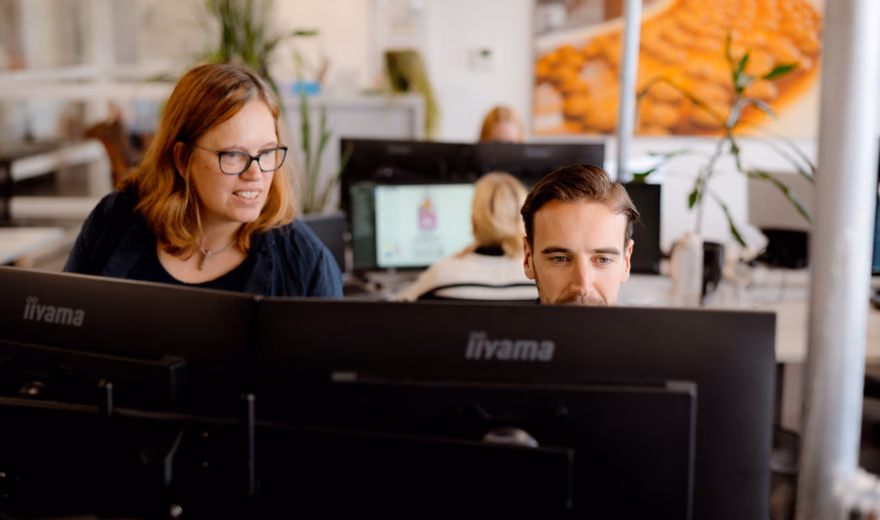 Twee medewerkers werken samen achter meerdere computerschermen in een moderne kantooromgeving.