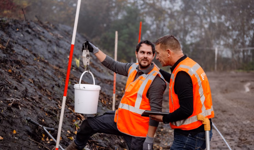 Men wearing orange reflective vests are inspecting a pile of soil, holding a bucket.