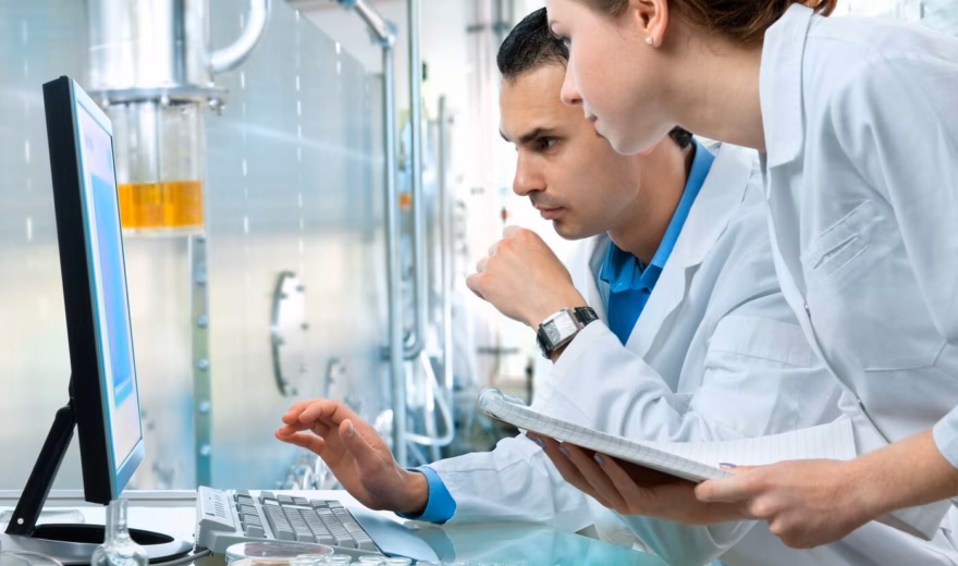 A man and a woman wearing white coats are reviewing information on a computer in a laboratory setting, with the woman holding a notebook.