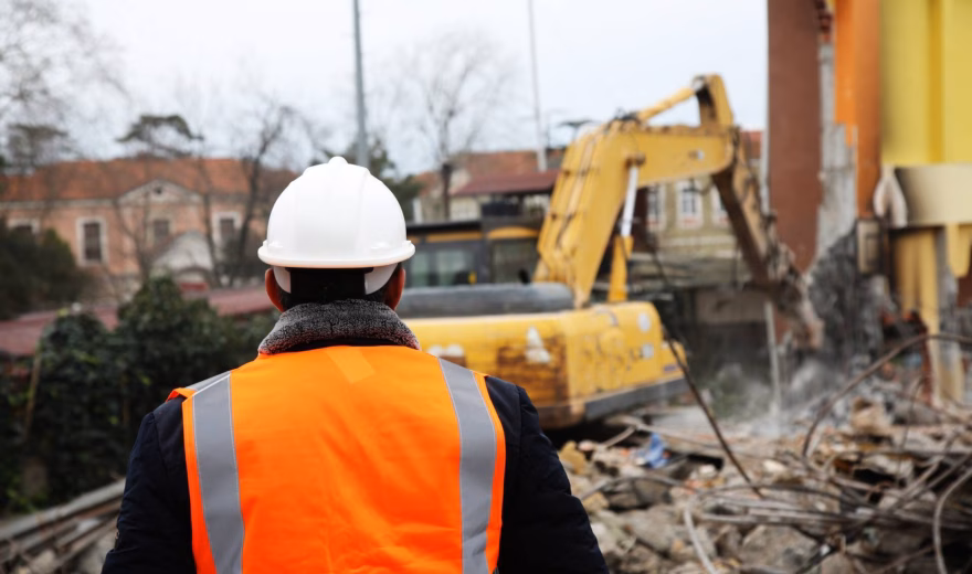 A person wearing a safety helmet and an orange safety vest is supervising demolition work being carried out with an excavator.