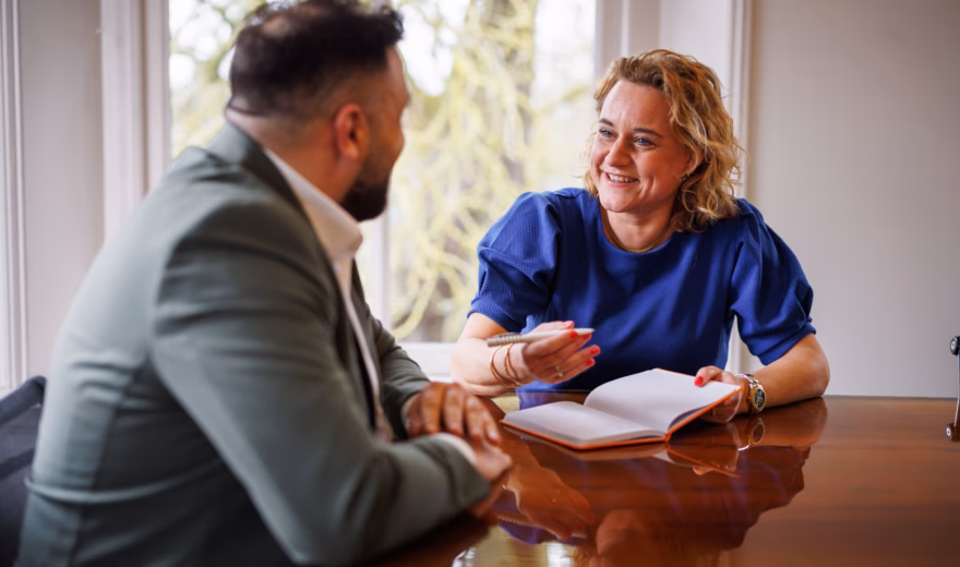 Man and woman in a meeting, with the woman wearing a blue top taking notes while smiling during the conversation.