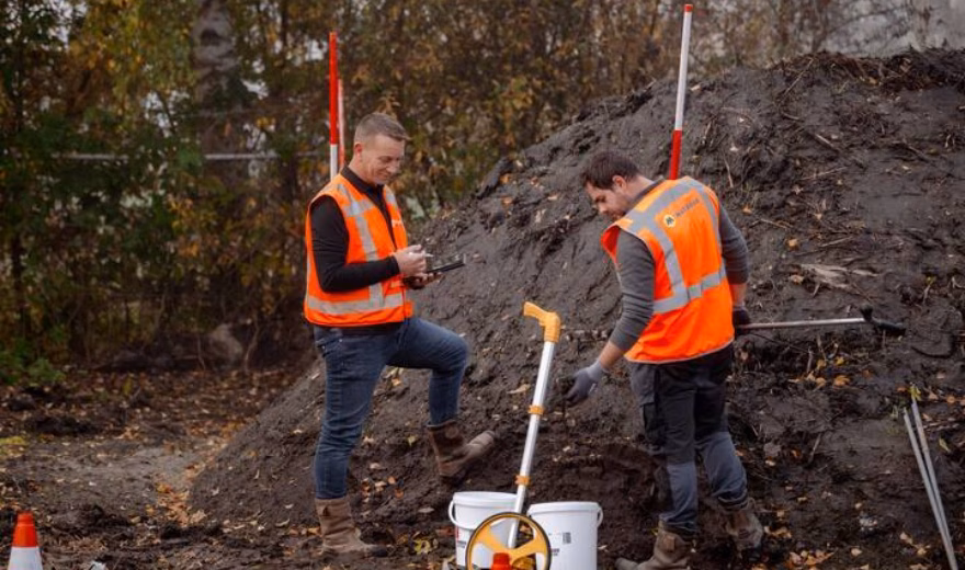 Two people wearing reflective vests inspecting a pile of soil, pointing at white buckets while one person takes notes on an iPad.