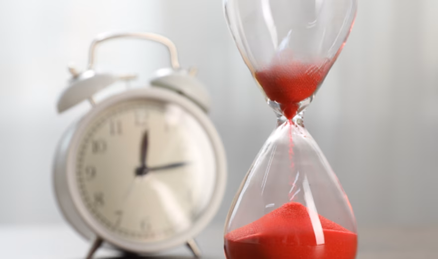 Im Vordergrund steht neben einem klassischen Wecker eine Sanduhr mit rotem Sand, die für schnelle Bearbeitungszeiten, effiziente Dienstleistungserbringung, Terminmanagement, zeitkritische Projekte und reaktionsschnellen Support bei Prüf-, Inspektions-, Zertifizierungs- und Konformitätsaktivitäten steht.