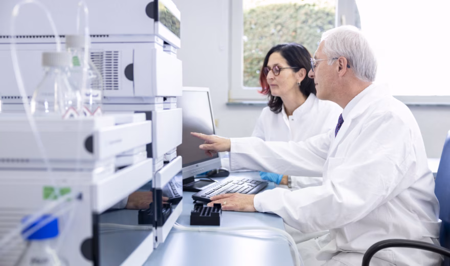 Zwei Labortechniker überprüfen Analysedaten an einem Computerarbeitsplatz, der sich in unmittelbarer Nähe modernster Laborgeräte befindet. Dieses Bild veranschaulicht wissenschaftliche Zusammenarbeit, Forschung, Qualitätskontrolle und Genauigkeitsprüfungen in einem modernen Labor.