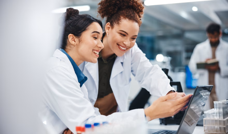 Two women in lab coats are smiling as they look at a laptop in a lab; scientific equipment can be seen nearby.