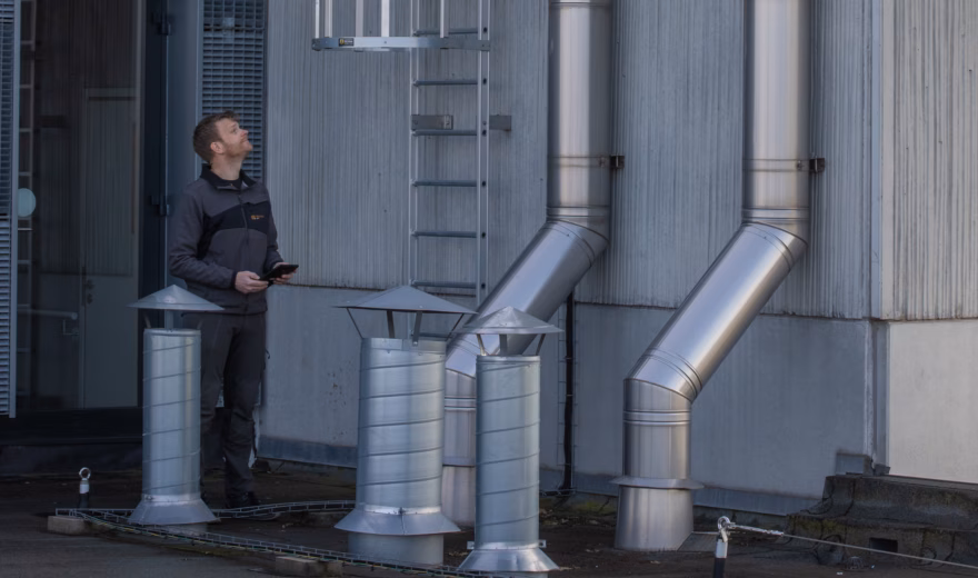 Un technicien inspecte les conduits de ventilation industriels et l'échelle de sécurité dans le cadre de l'accompagnement du CO‑Covenant et du contrôle des flux d'air et des émissions.
