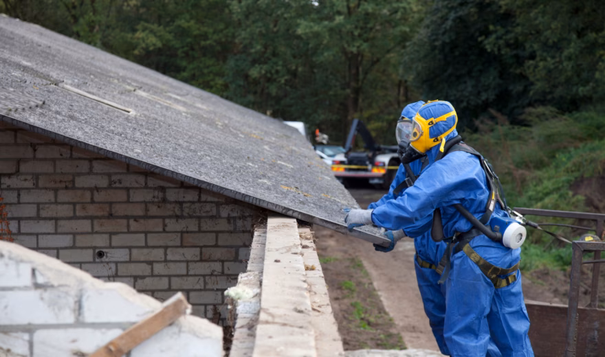 Deux employés vêtus de combinaisons de protection intégrales effectuent une inspection de l'amiante sur une dalle de toiture en extérieur.
