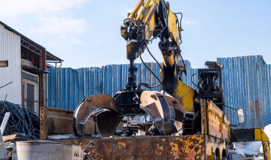 Une grue de chargement sur camion équipée d'une pince déplace des matériaux dans une zone industrielle.