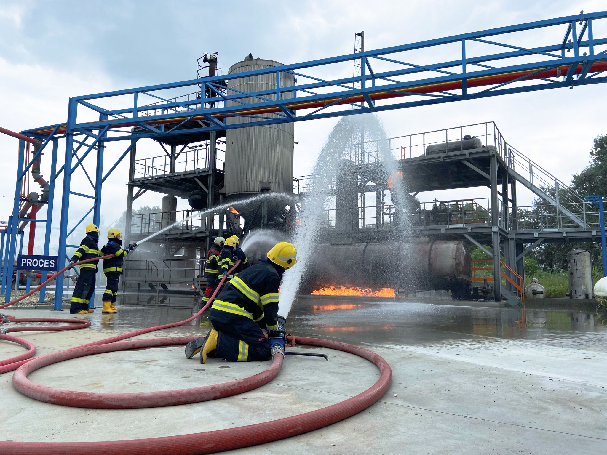 Feuerwehrleute führen an einer Industrieanlage eine Löschübung mit sichtbarem Feuer und Löschwasser durch.