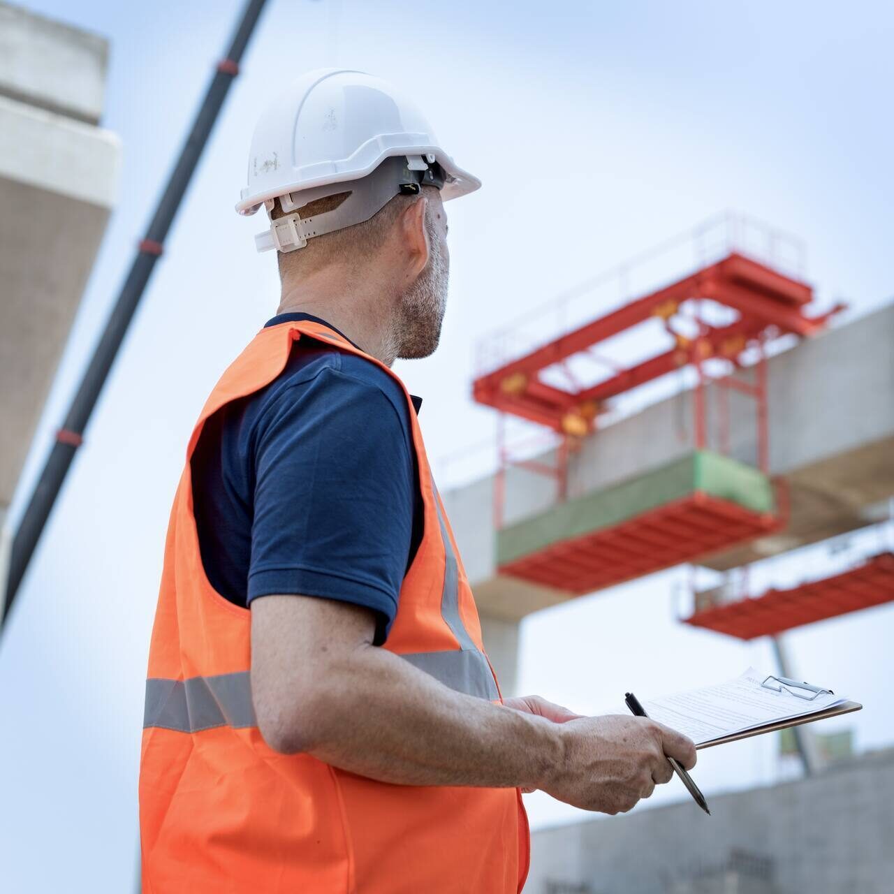 A person wearing a safety helmet and an orange safety vest at a construction site, with a building structure visible in the background.
